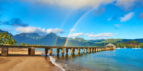 the beautiful Hanalei Bay Pier in the morning with a double rainbow over the pier and the lush towering mountains of Hanalei Kauai in the background