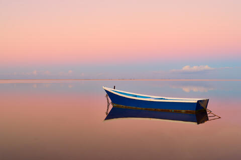 A solitary blue boat rests on perfectly still water at sunrise, surrounded by soft pastel hues of pink and blue reflecting across the sky and horizon, creating