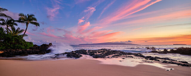 Vibrant pink and violet sunset clouds over the Maui coastline with ocean waves, palm trees, and volcanic rock at dusk.