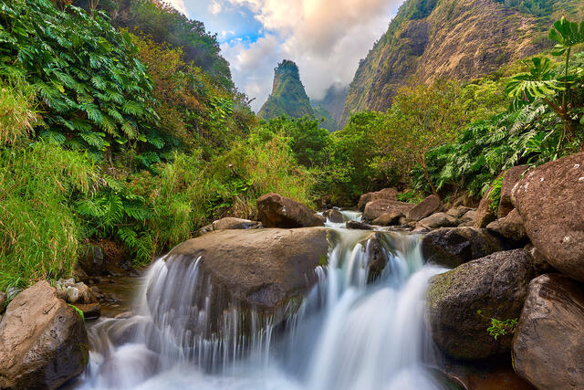 a waterfall flows over the rocks at Iao Valley State Park with the Iao Needle iconically in the background of the lush forest.  Hawaii Photography 