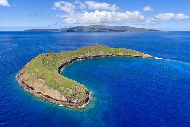 Aerial view of Molokini Crater off Maui, Hawaii, showing its crescent shape surrounded by deep blue ocean under a bright sky.