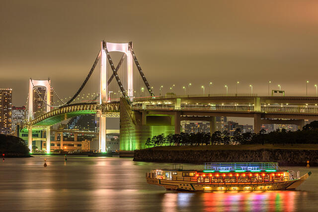 rainbow bridge in Tokyo, Japan illuminated at night with a colorful boat/taxi in the water