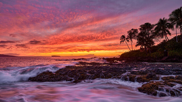 stunningly vibrant sunset at secret beach in Makena on the island of Maui with incoming waves over the lava rocks.  Photographed by Andrew Shoemaker