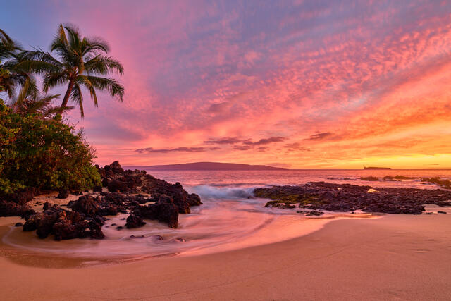 Secret Glow is a stunning beach sunset landscape fine art photograph featuring moving water, palm trees, rocks, and a stunning beach.