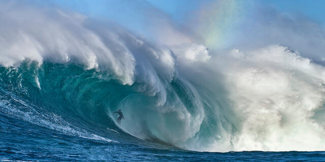 the biggest wave in Hawaii Jaws also known at Peahi with surfer Kai Lenny in the barrel and a rainbow visible from the spray.  Photographed by Andrew Shoemaker