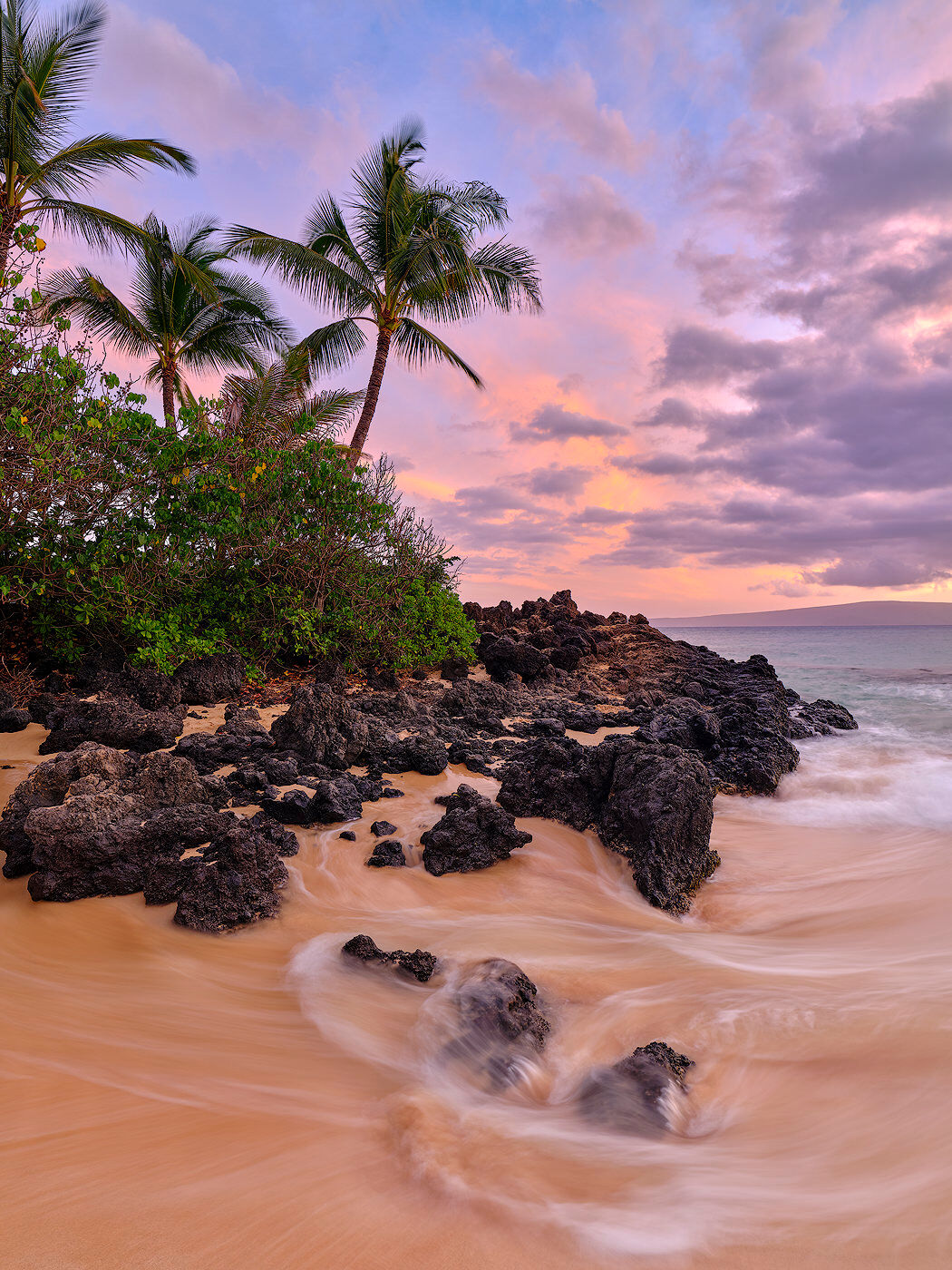 A beautiful vertical photo with silky water, coconut palm trees and a colorful pastel sunset at Secret Beach in Makena.  Photographed by Andrew Shoemaker