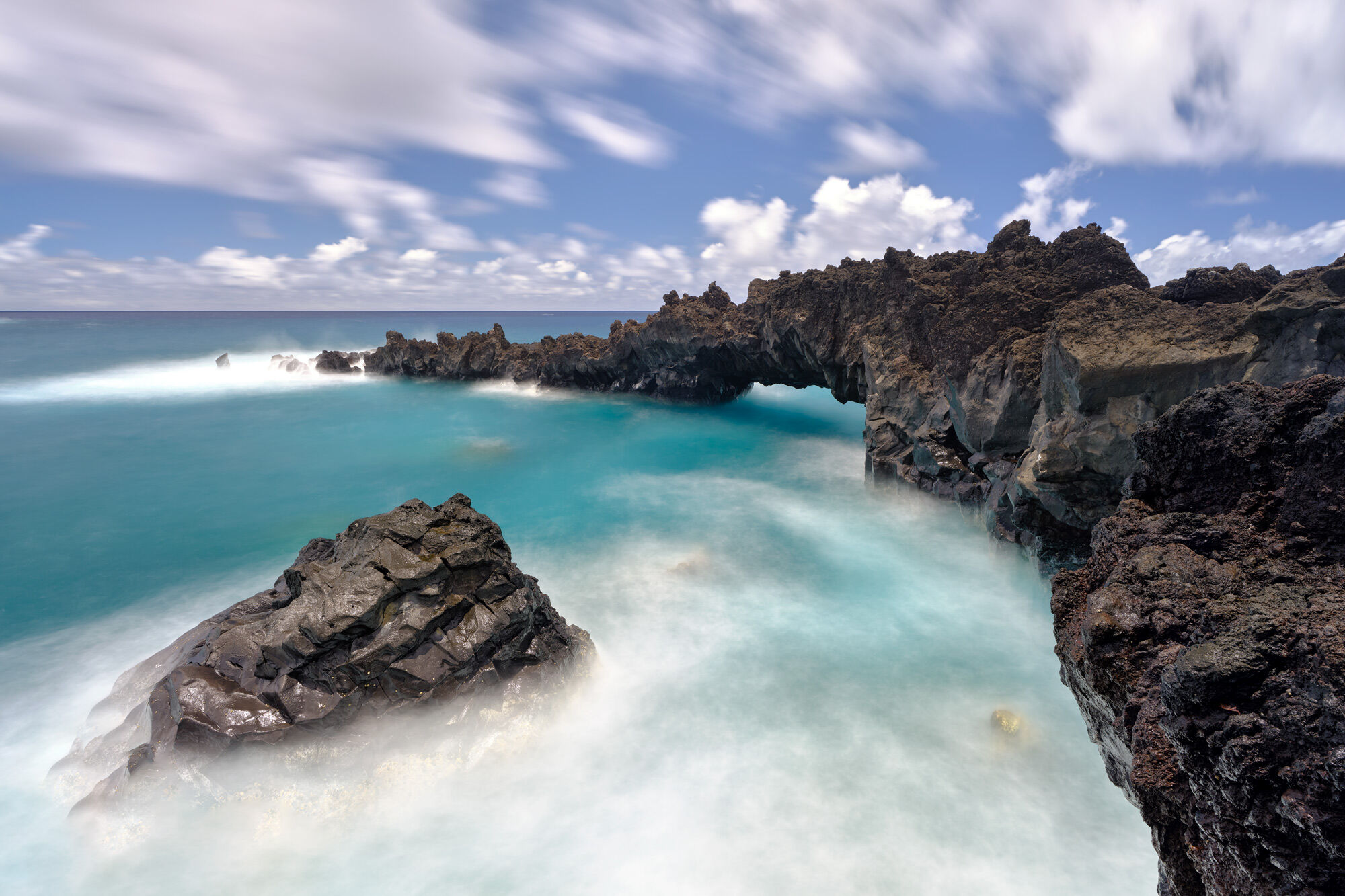 a long exposure captured of an arch made from lava rock showcasing beautiful aqua colored water and streaking clouds by Andrew Shoemaker