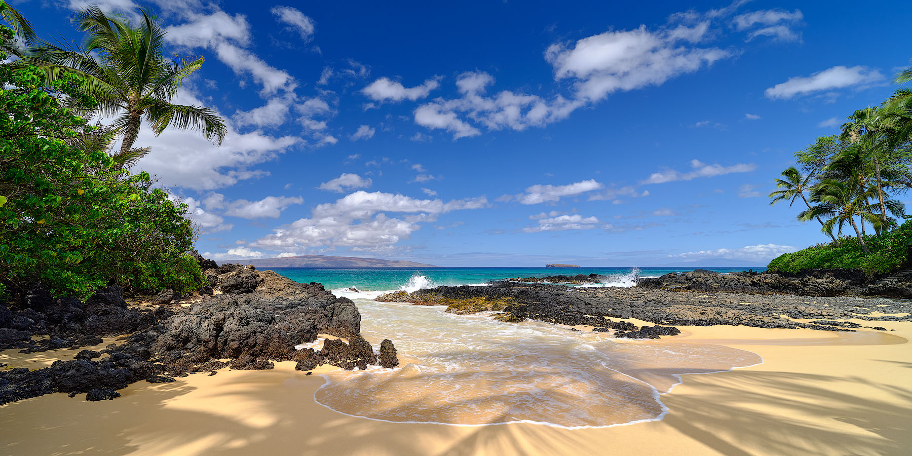 panoramic view of Secret Beach on the island of Maui with the shadows from the palm trees in the foreground as a wave comes in