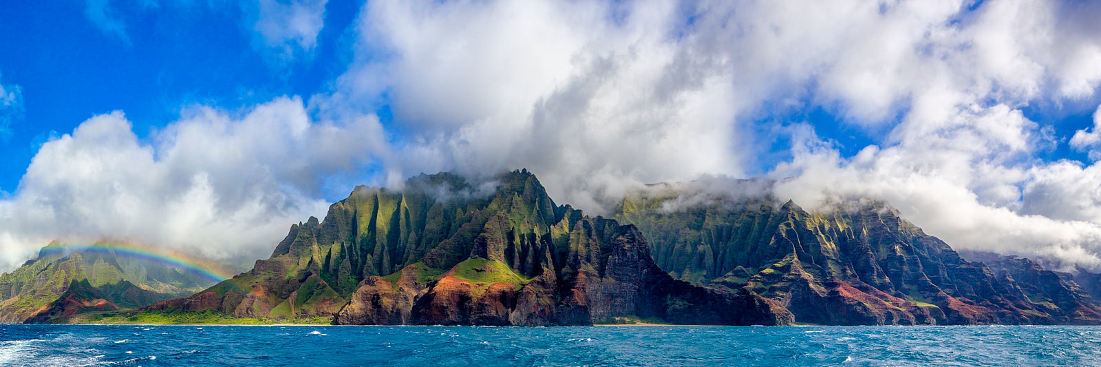 panoramic photo of the Na Pali coastline on the Hawaiian island of Kauai featuring a rainbow, dramatic clouds, and vibrant blue water