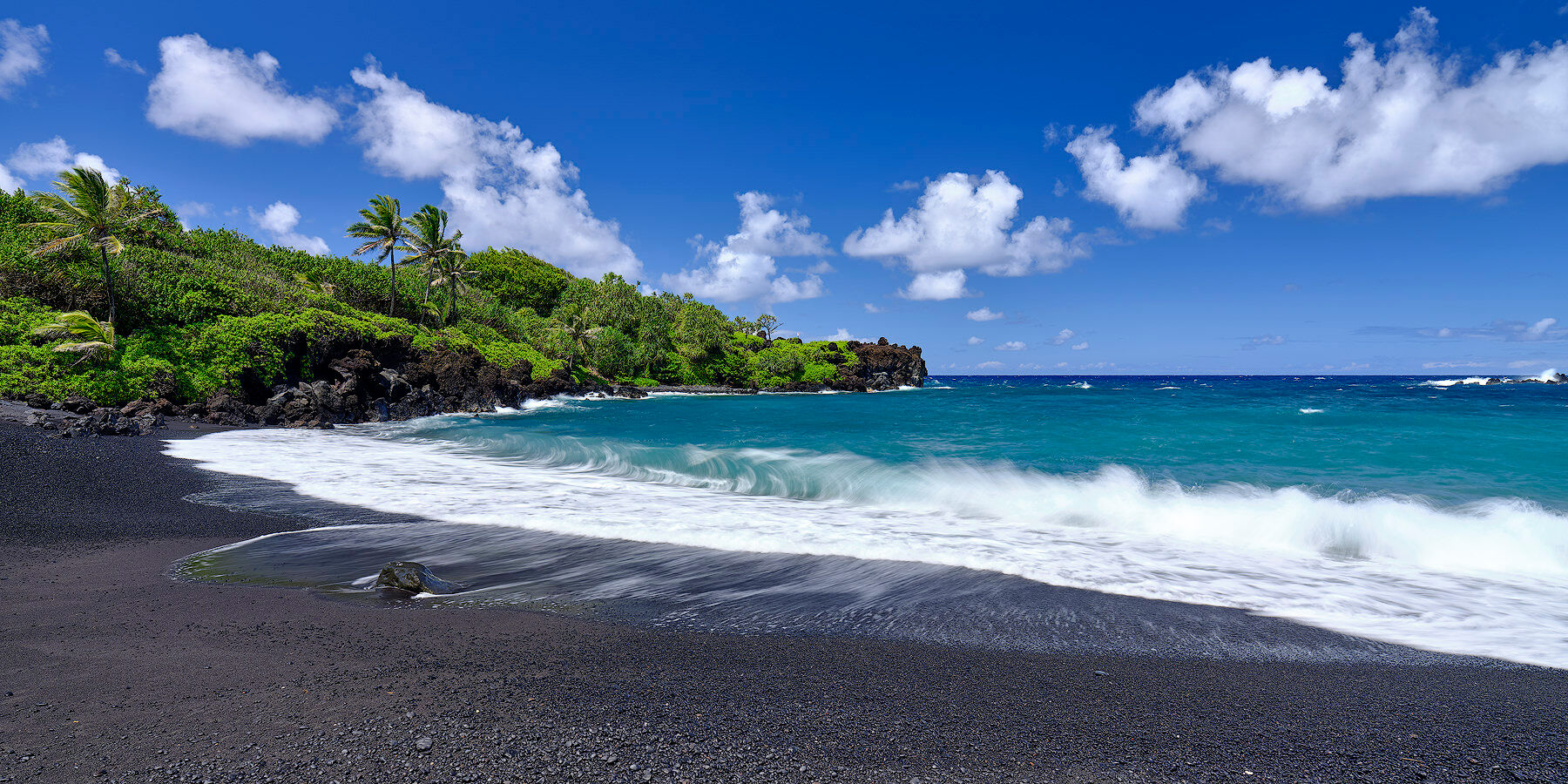 panoramic daytime view of the black sand beach at Waianapanapa State Park near Hana on the island of Maui with an incoming wave